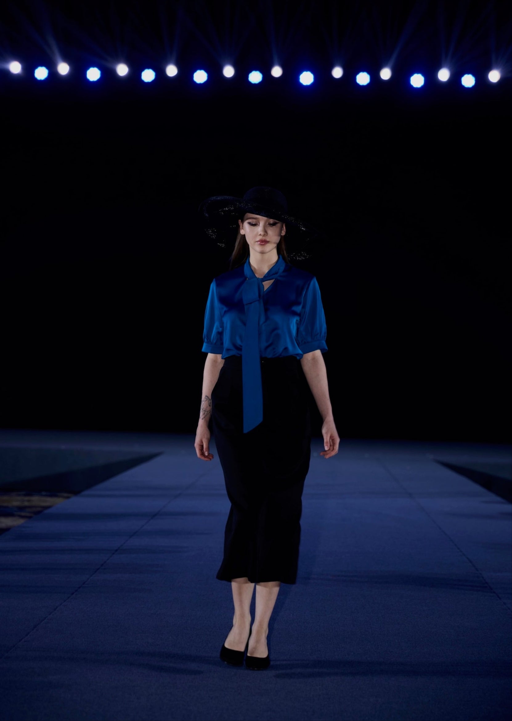 Model wearing a blue blouse and black skirt on a runway with dark background and blue lights.
