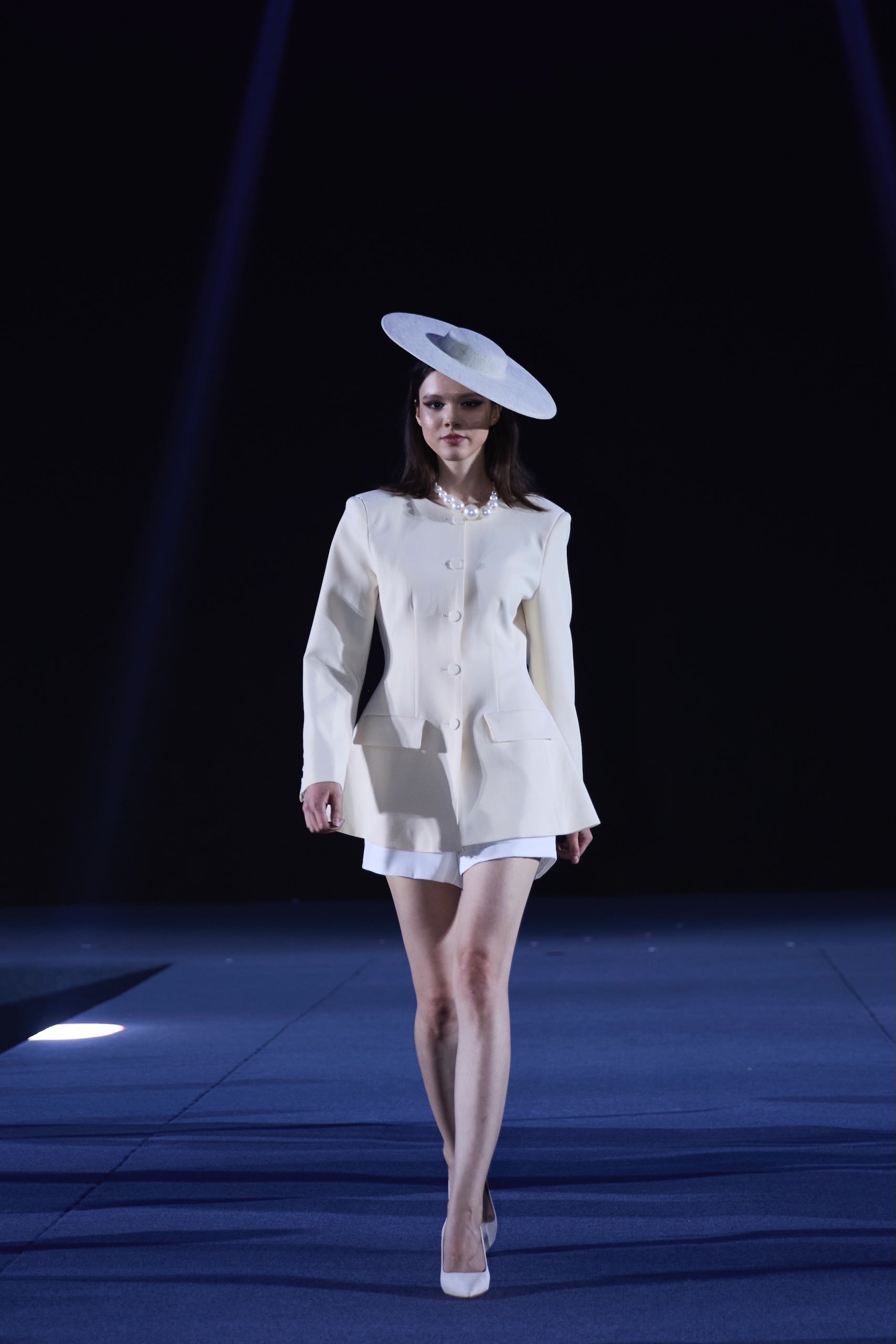 Model wearing a white outfit with a large hat on a runway.