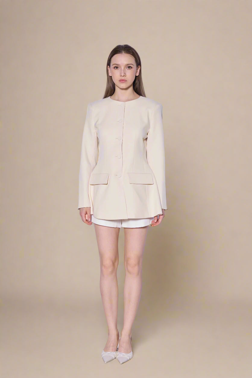 Woman wearing a beige blazer and white shorts against a white curtain background