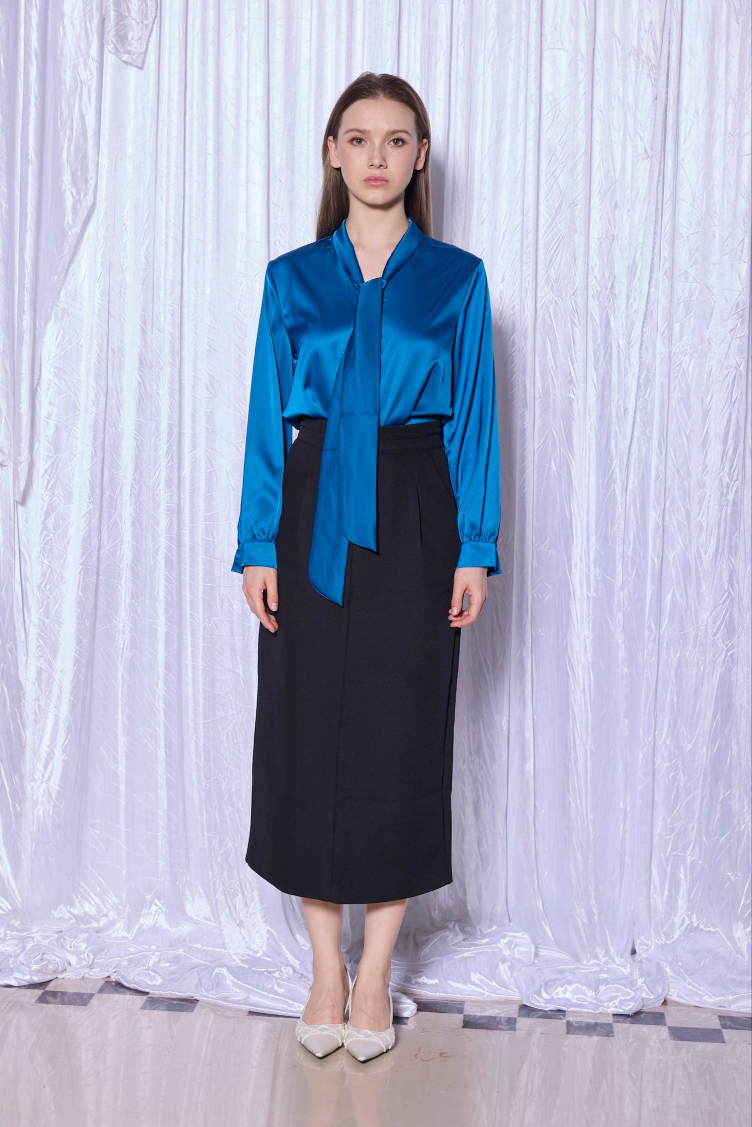 Woman wearing a blue satin blouse and black skirt against a white curtain background