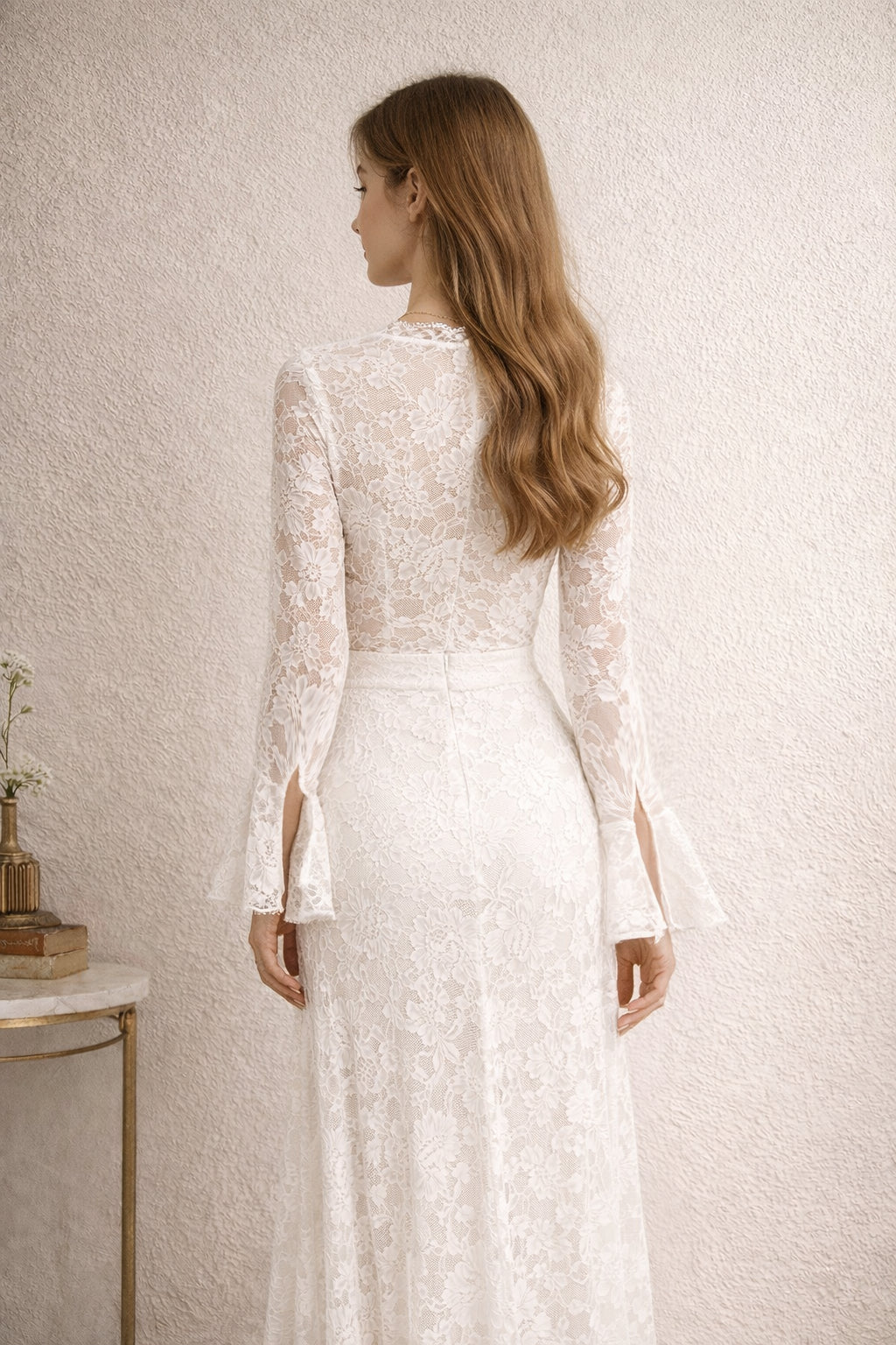 Woman wearing a white lace dress standing against a plain wall.