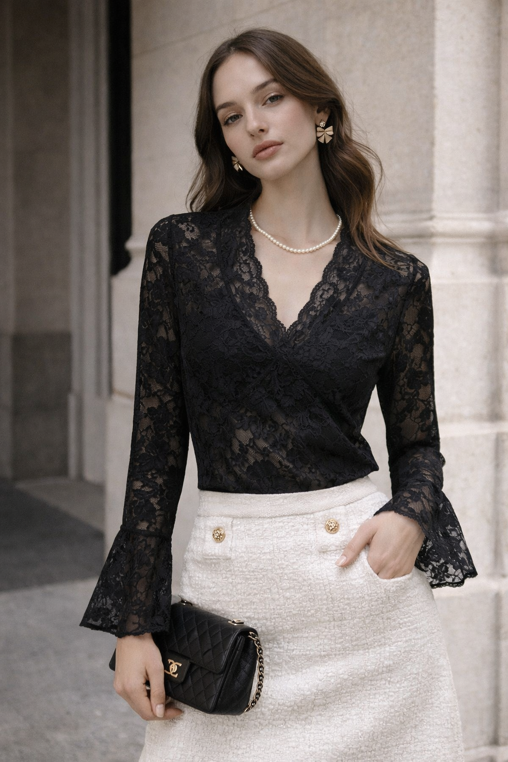 Woman wearing a black lace top and white skirt with a black handbag, standing against a neutral background.
