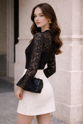 Woman wearing a black lace top and white skirt with a black clutch against a stone wall.