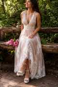 A woman wearing a sleeveless lace and tulle high-low dress sits on a rustic wooden bench outdoors, with pink flowers beside her and a wooded background.
